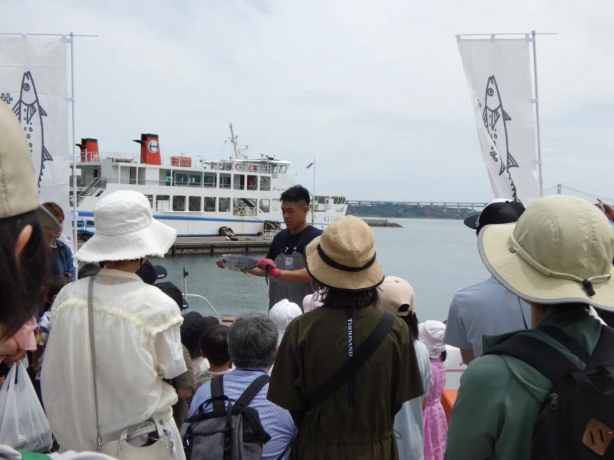 港で、作業着を着た男性が捕れたての魚を掲げ、多くの見物人がそれを見ている様子。フェリーや魚の旗が見え、地元のイベントのようだ。