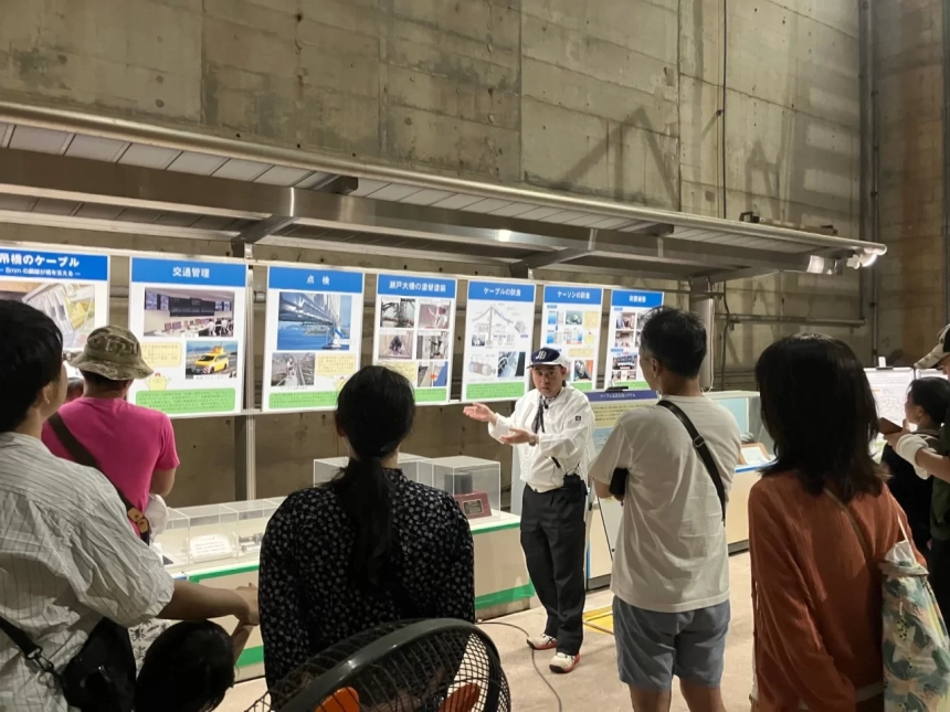 瀬戸大橋に関する展示会場で、講師が来場者に向けて橋の交通管理、点検、メンテナンスなどについて説明している様子です。複数のパネルが設置され、人々が熱心に耳を傾けています。