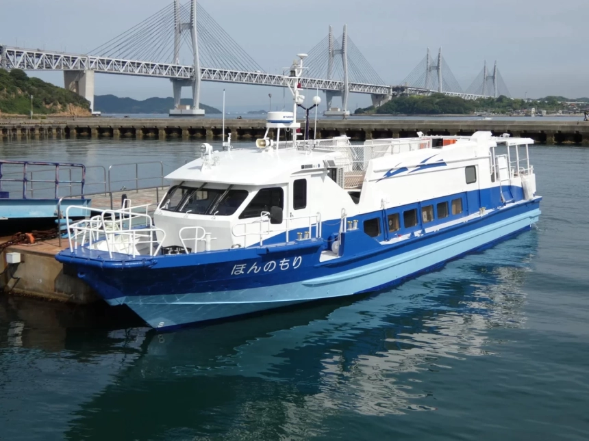 青と白の船が港に停泊しており、その背景には雄大な吊り橋と島々が広がっています。穏やかな水面に船体が映り込み、のどかな港の風景を捉えています。